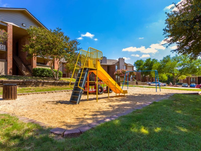 Valley Creek Playground with a yellow slide, swing set, trees, and apartment buildings in the background on a sunny day.