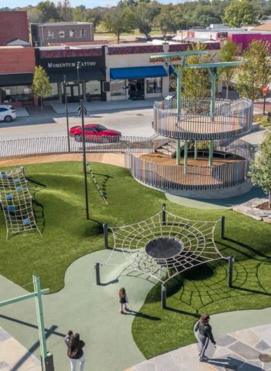 Valley Creek Aerial view of a playground with climbing nets and tower, surrounded by shops and trees on a sunny day.