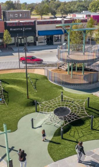 Valley Creek Aerial view of a playground with climbing nets and tower, surrounded by shops and trees on a sunny day.